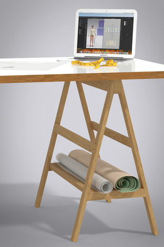 Design Desk scene of 3D objects. Design desk has white laminate top and wood sides with wooden saw horse legs. Scene showing corner of desk with laptop, tape measure and a roll of pattern paper and oaktag paper on the shelf of the saw horse legs. 