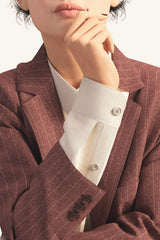 Female avatar in digital mauve suit jacket with white pinstripe. Avatar has right hand up by mouth and elbow held by left hand. Wearing cream button down shirt under jacket, showing sleeve cuff details of both jacket and shirt. Cropped just above lips and just above elbow at bottom.